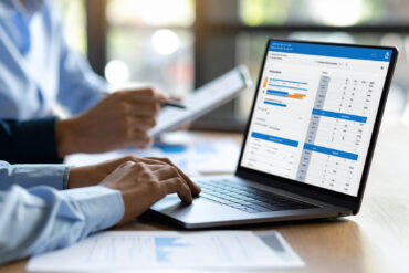 Businessman working laptop with financial data and charts, office environment with natural light, hands typing keyboard, blurred background with colleague holding tablet, teamwork and analysis