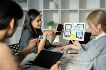 Diverse team collaborating on UX UI design, using digital devices and wireframes in a modern office setting.